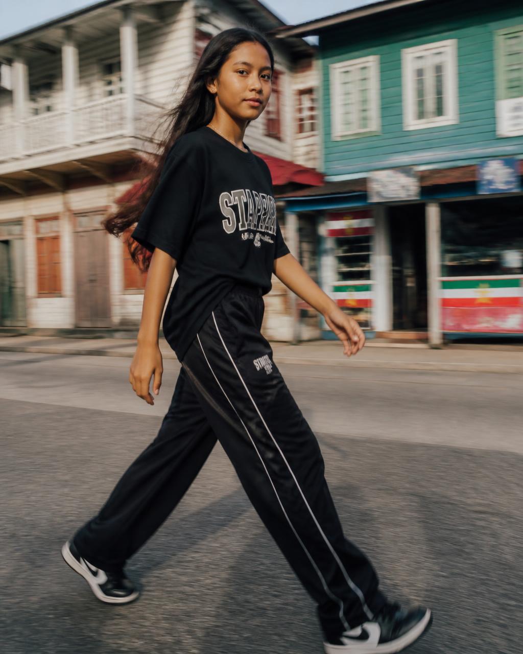 Girl in a black FAKA tee and track pants walking a colorful Paramaribo street